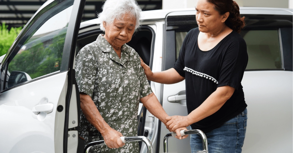 A female care worker assists an elderly woman holding a mobility walker out of a van.