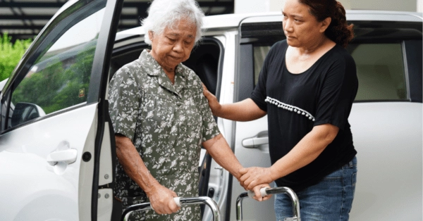 A female care worker assists an elderly woman holding a mobility walker out of a van.
