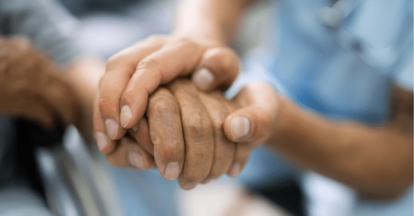Nurse holds hand of elderly patient. Focus of image is on the hands held, with the patient and nurse out of focus in the background.