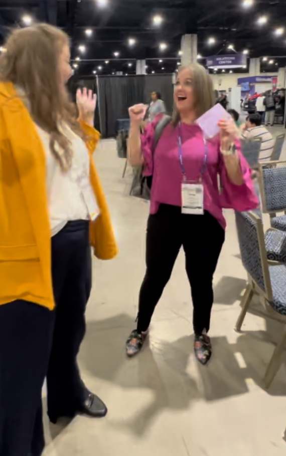 A woman in a yellow coat hands a gift card to an excited woman in a pink shirt