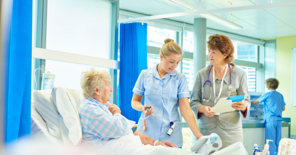 Two female medical professionals talk to elderly patient in hospital bed.