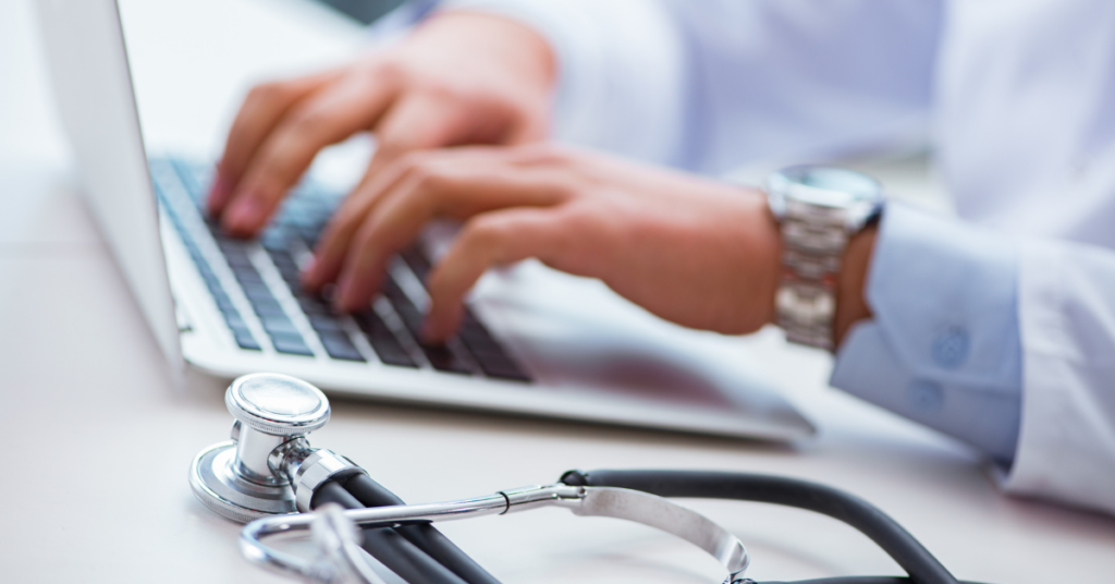 Doctors hands in view as they type on a laptop. Stethoscope is on table in front of laptop
