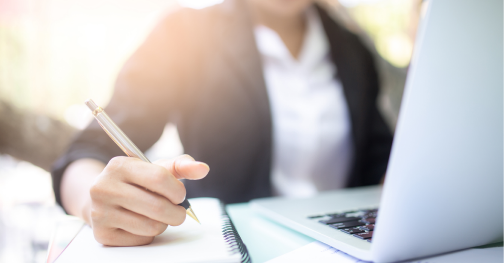 Woman in suit writing on piece of paper with a pen. In the foreground, she is sitting behind a laptop