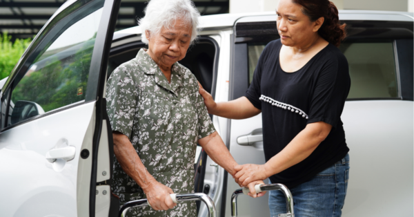 A female care worker assists an elderly woman holding a mobility walker out of a van.