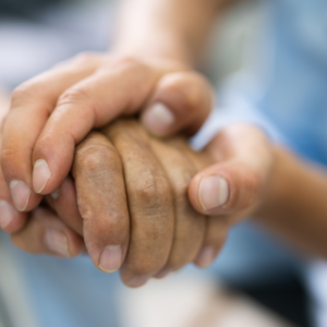 Nurse holds hand of elderly patient. Focus of image is on the hands held, with the patient and nurse out of focus in the background.